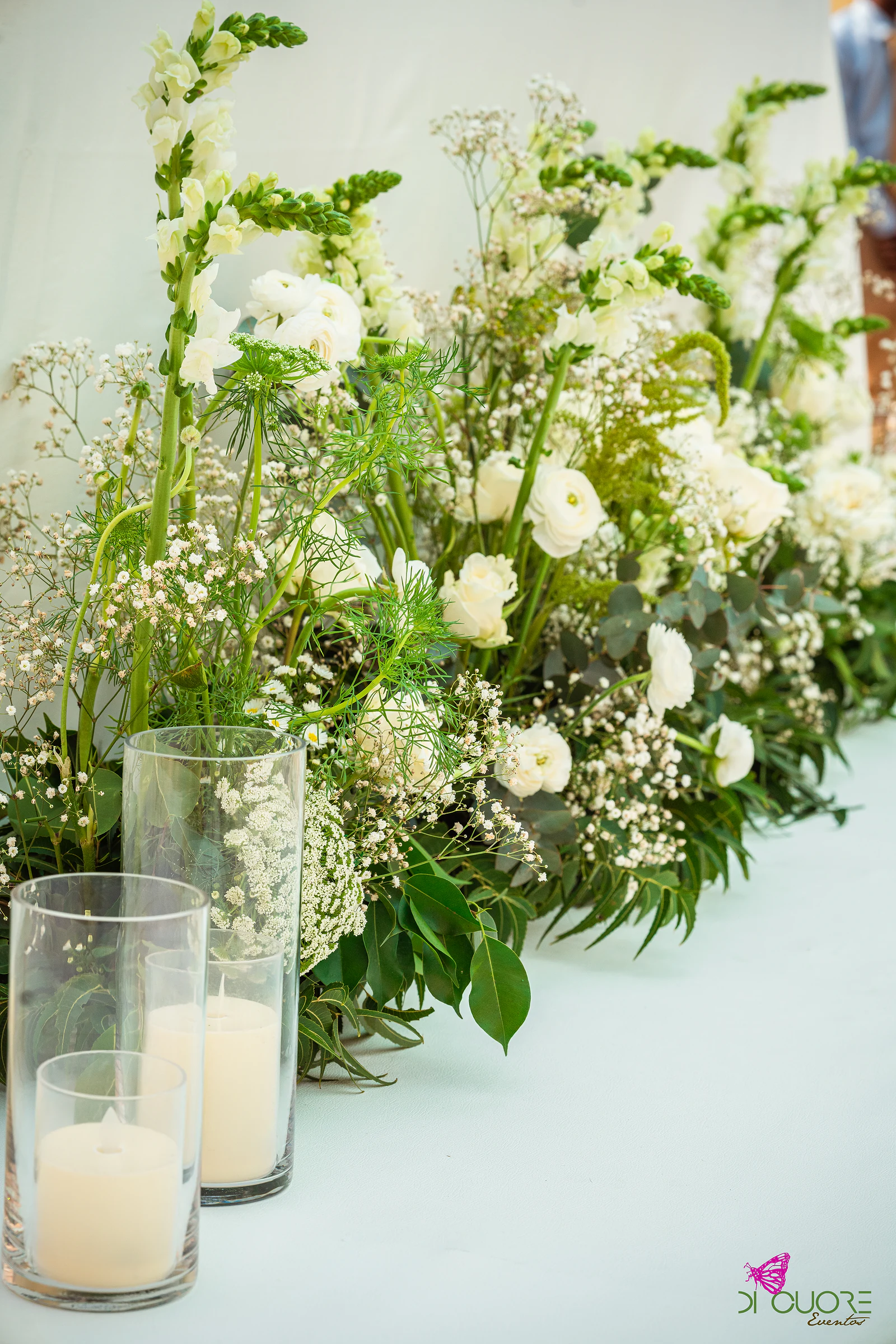 Arranjo baixo de flores brancas e folhagem com velas em cilindros de vidro, sobre uma superfície branca