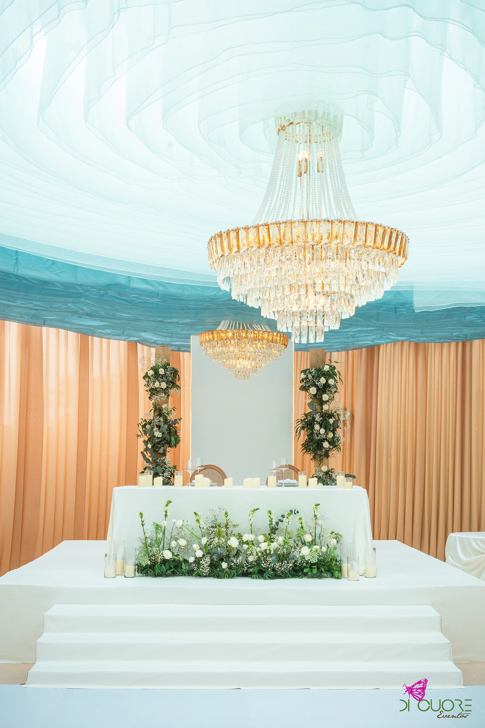 Mesa dos noivos sobre degraus brancos, com flores brancas e verdes, sob um lustre de cristal e um tecto branco e azul-turquesa