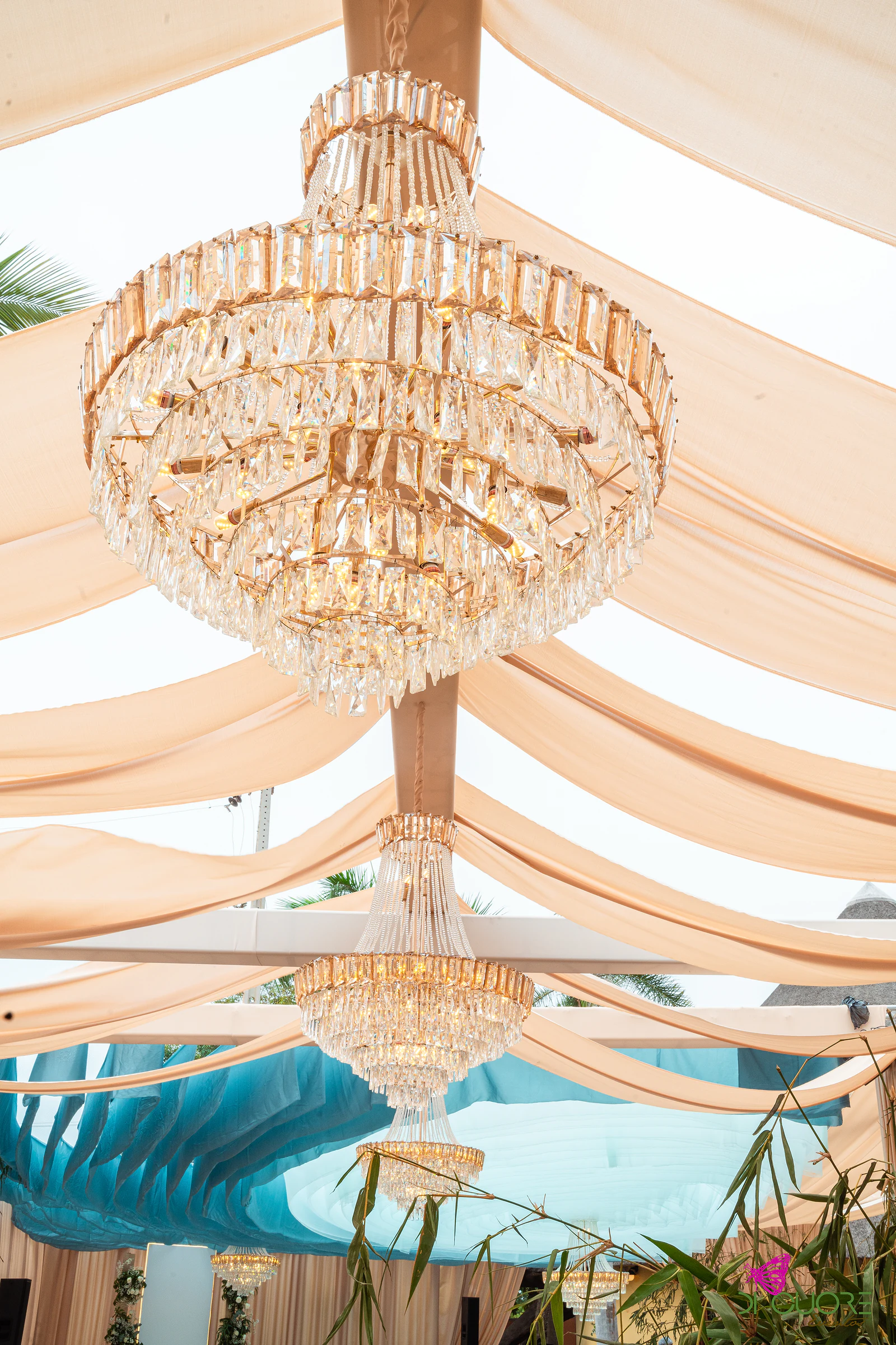 Lustre de cristal dourado suspenso entre tecidos cor de pêssego, com outros lustres e o tecto azul-turquesa ao fundo