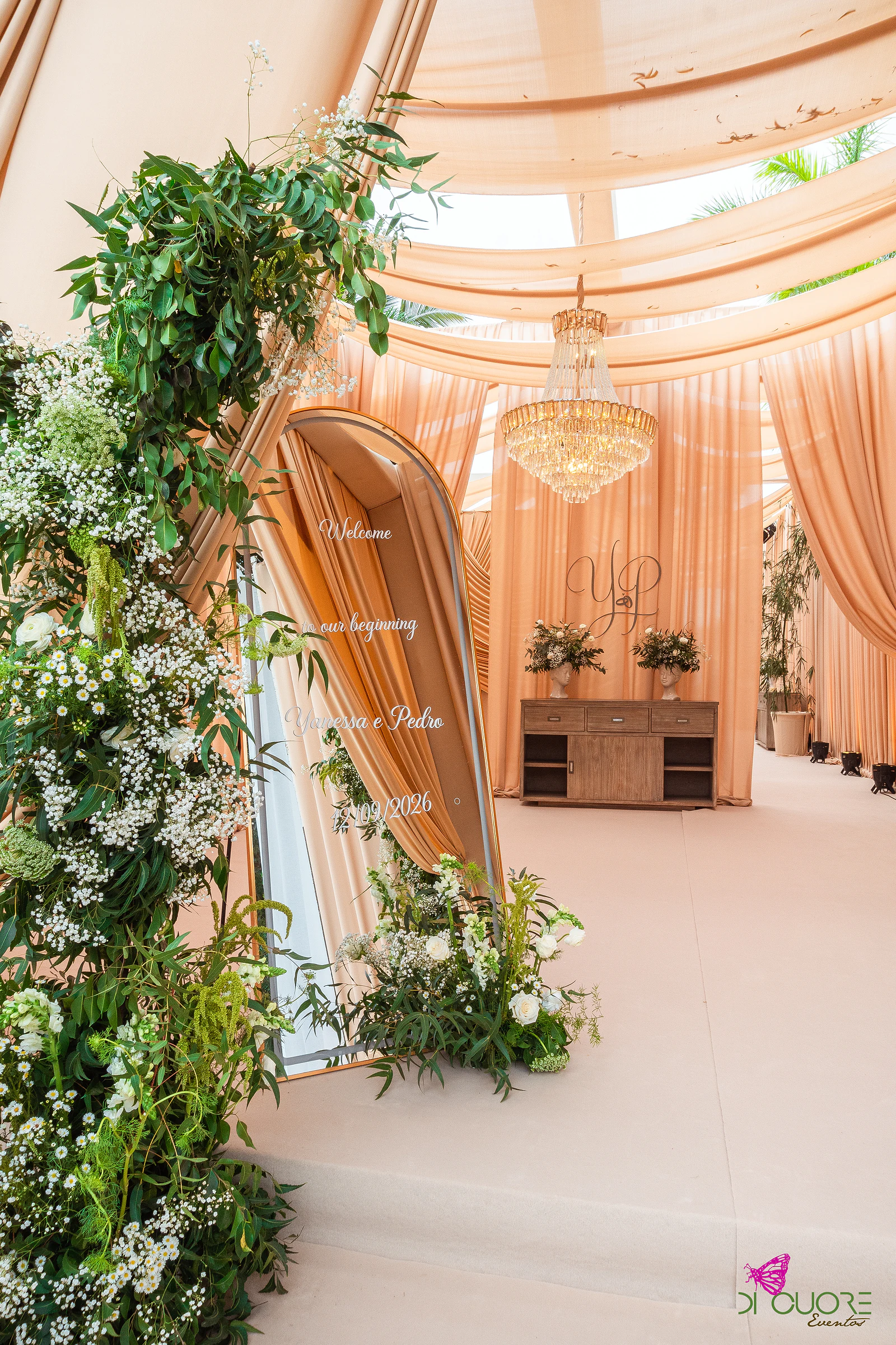 Sala com um arco de folhagem e flores brancas miúdas à esquerda, um painel de boas-vindas em arco, uma cómoda de madeira ao fundo e um lustre de cristal sob cortinados cor de pêssego