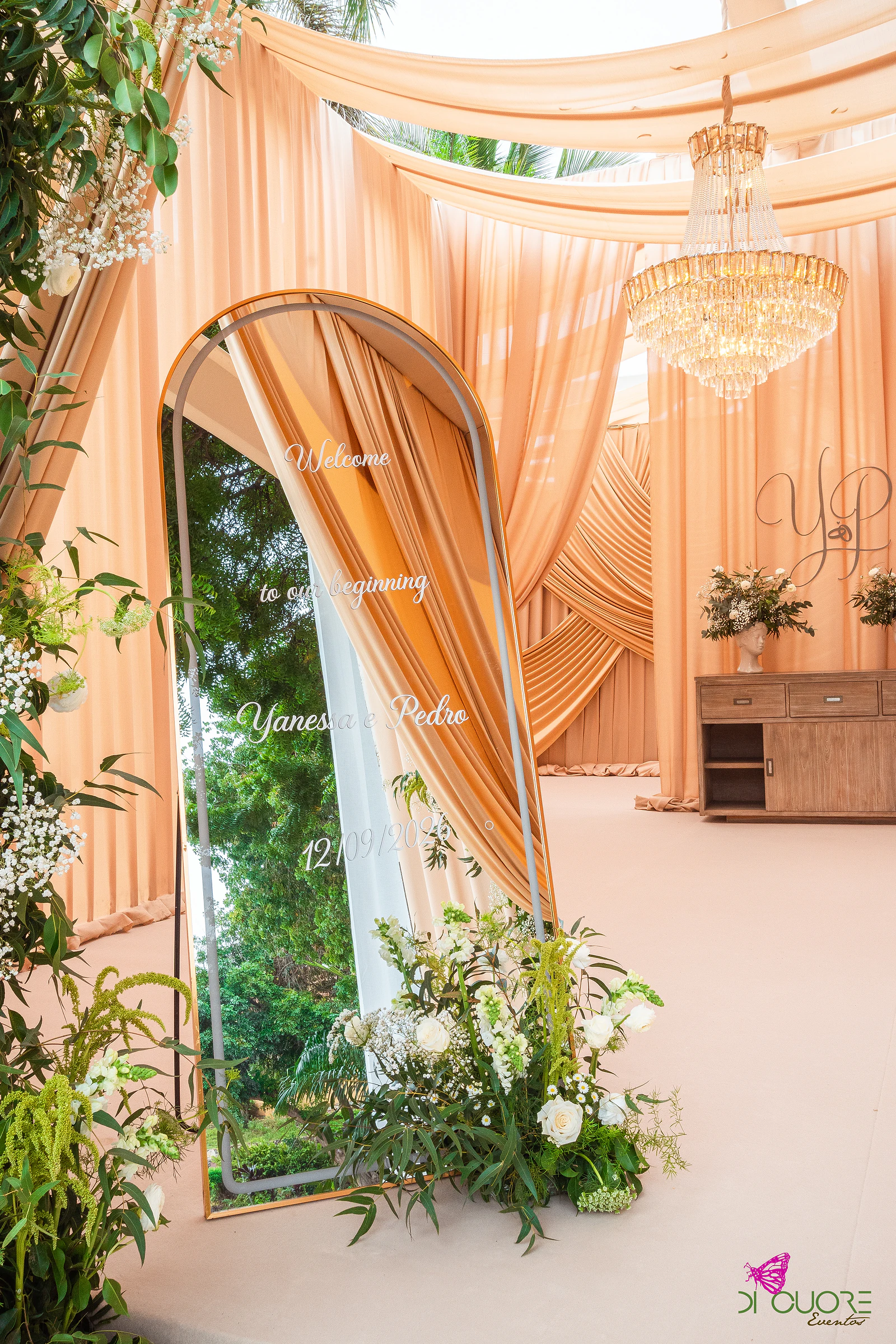 Espelho em arco com mensagem de boas-vindas, junto a um arranjo de flores brancas e folhagem, com um aparador sob um lustre e cortinados cor de pêssego ao fundo