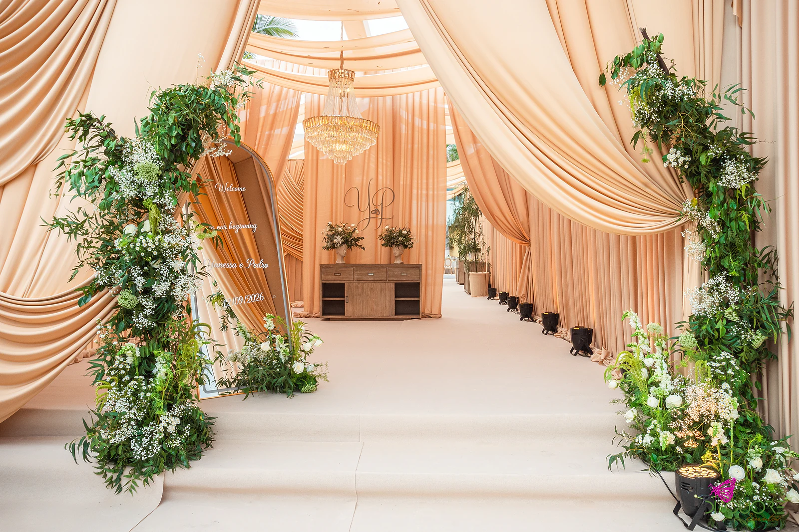 Corredor de entrada entre cortinados cor de pêssego, com um arco de folhagem e flores brancas junto ao espelho de boas-vindas e um aparador ao fundo