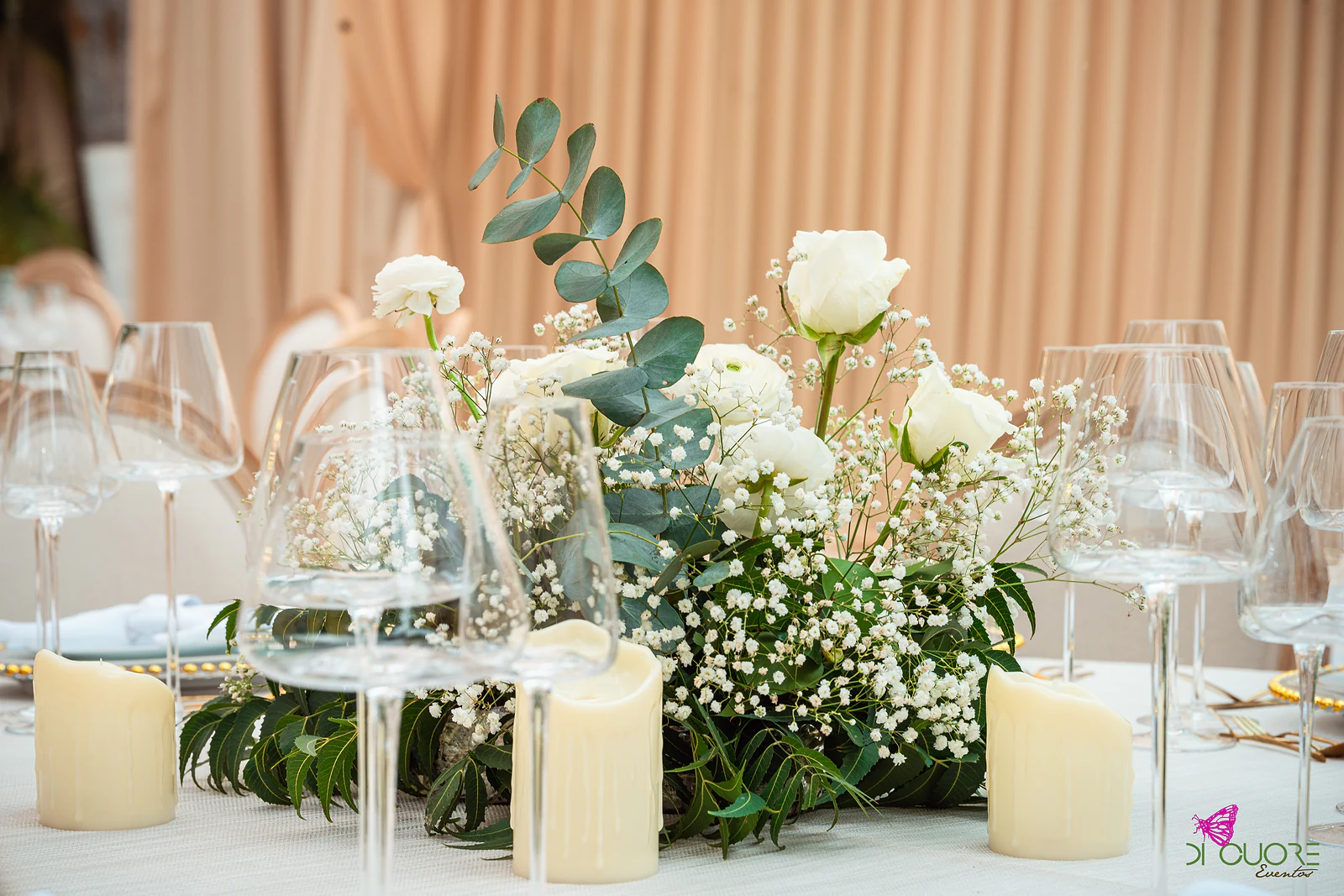 Centro de mesa baixo com rosas brancas, gipsófila e eucalipto, entre velas e copos de cristal