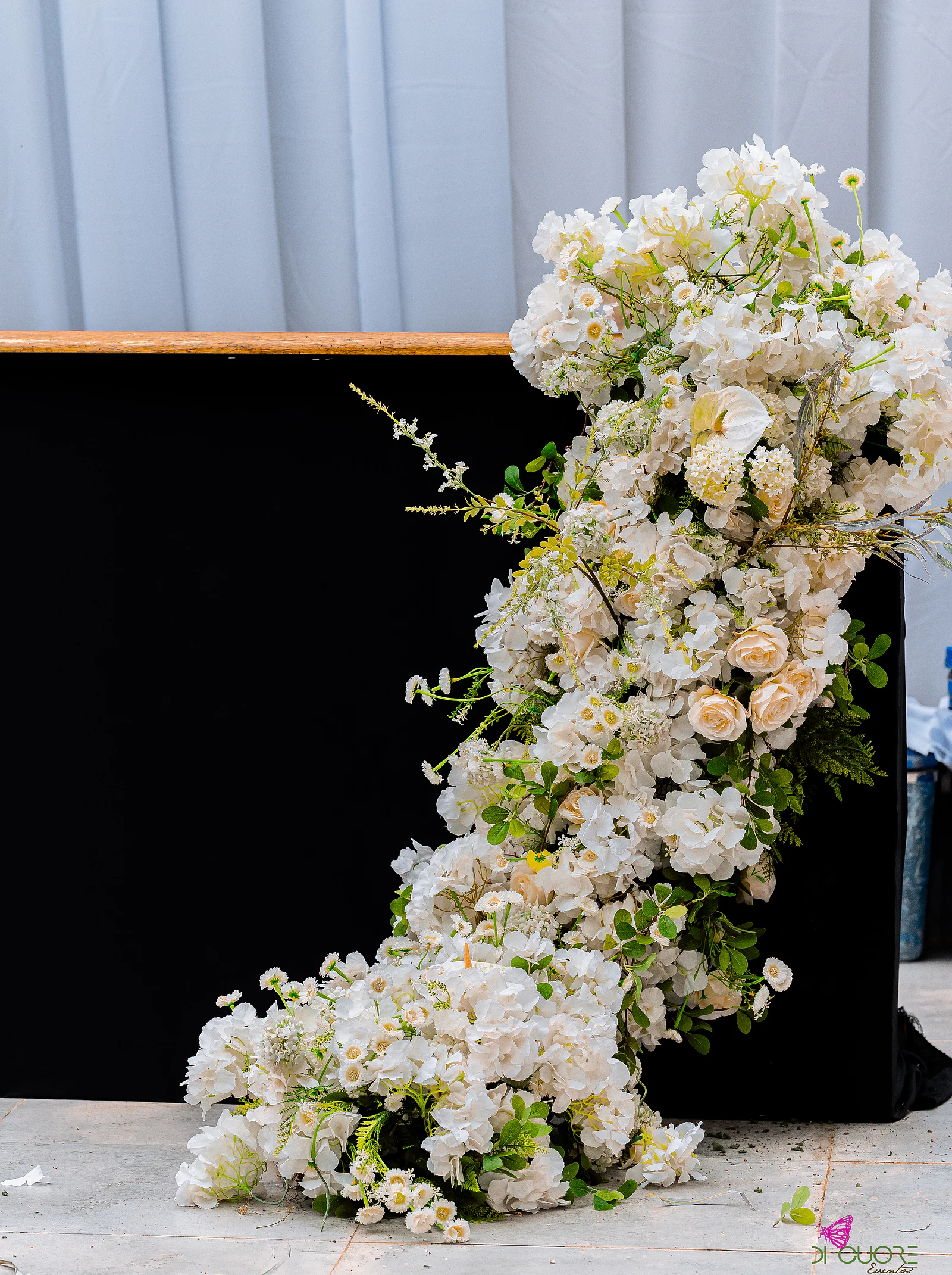 Cascata de flores brancas, rosas e antúrio sobre um painel preto, diante de um cortinado claro