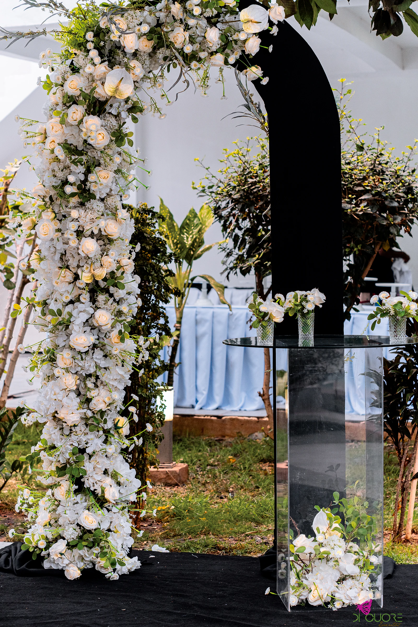 Arco de rosas e hortênsias brancas ao lado de um painel curvo preto e de um pedestal de acrílico com flores