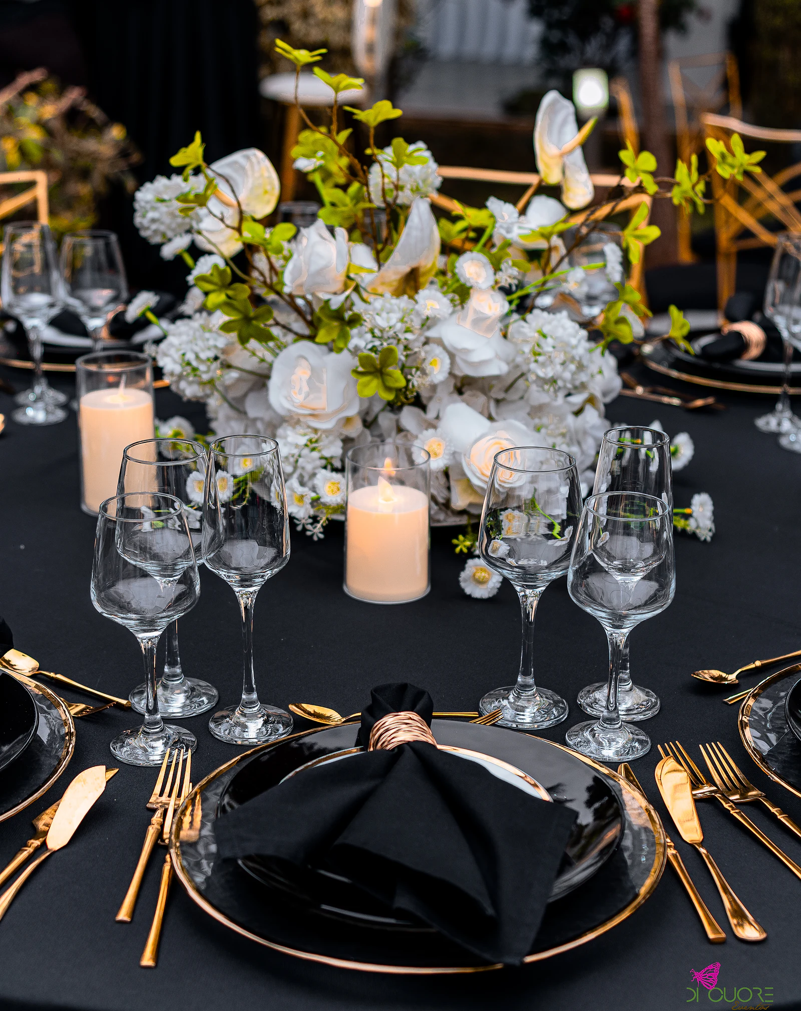 Mesa de toalha preta com prato negro de rebordo dourado, guardanapo preto, talheres dourados, copos de cristal, velas e centro de flores brancas