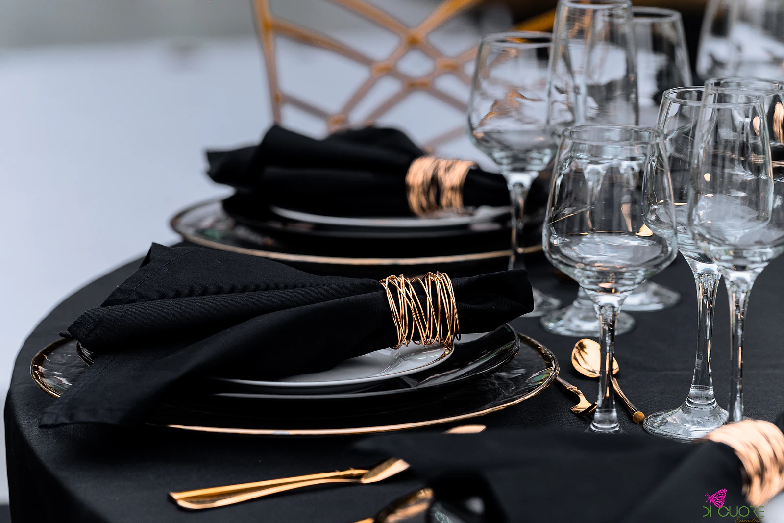 Prato negro de rebordo dourado com guardanapo preto preso por uma argola dourada, entre copos de cristal e talheres dourados