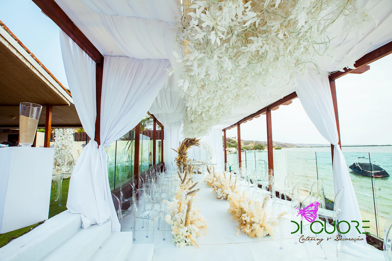 View along the ceremony table between white drapes and a cloud of hanging white leaves, with pampas plumes and the sea on the right