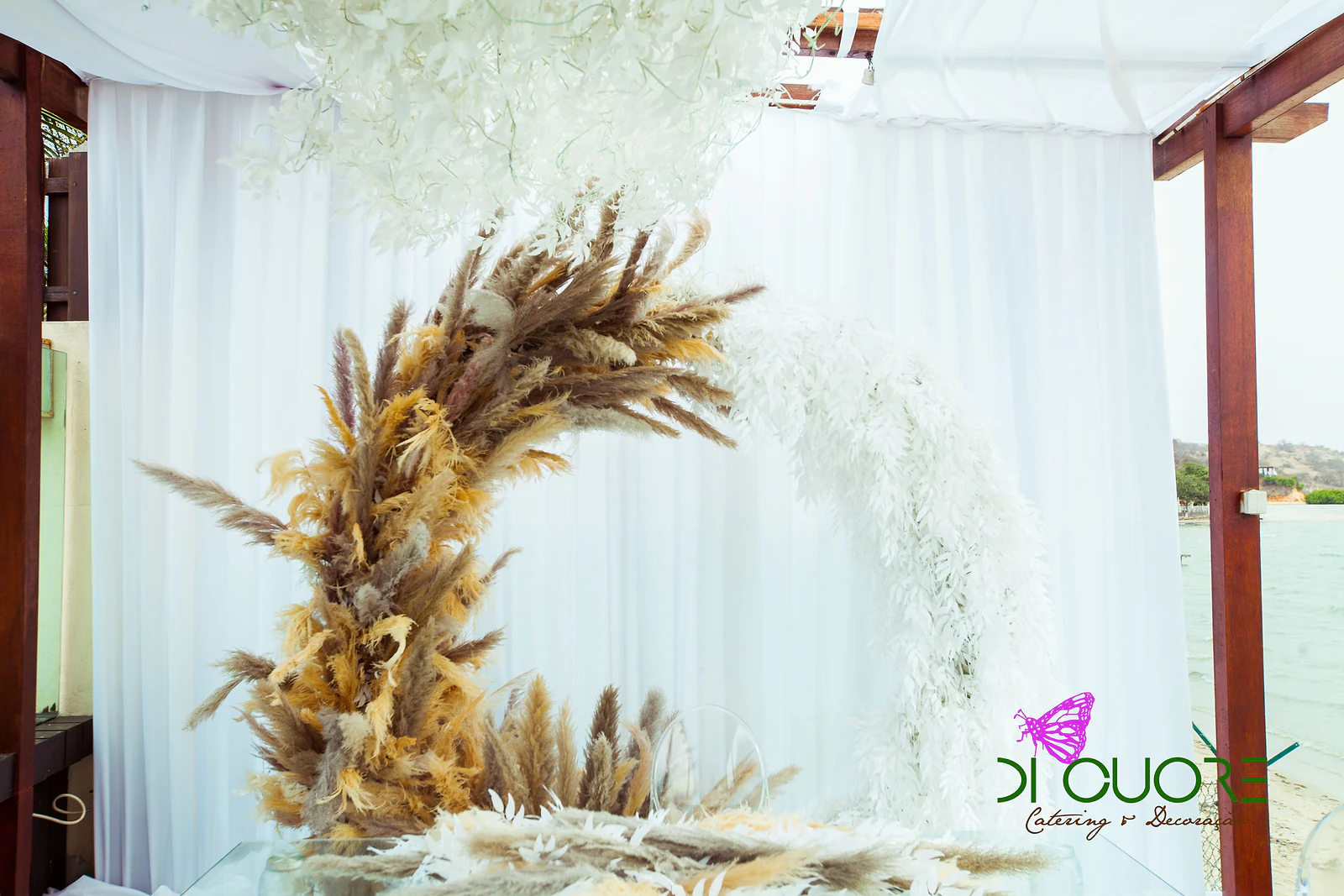 Half-moon arch, half plumes of pampas grass and half white leaves, in front of white drapes and a glass table