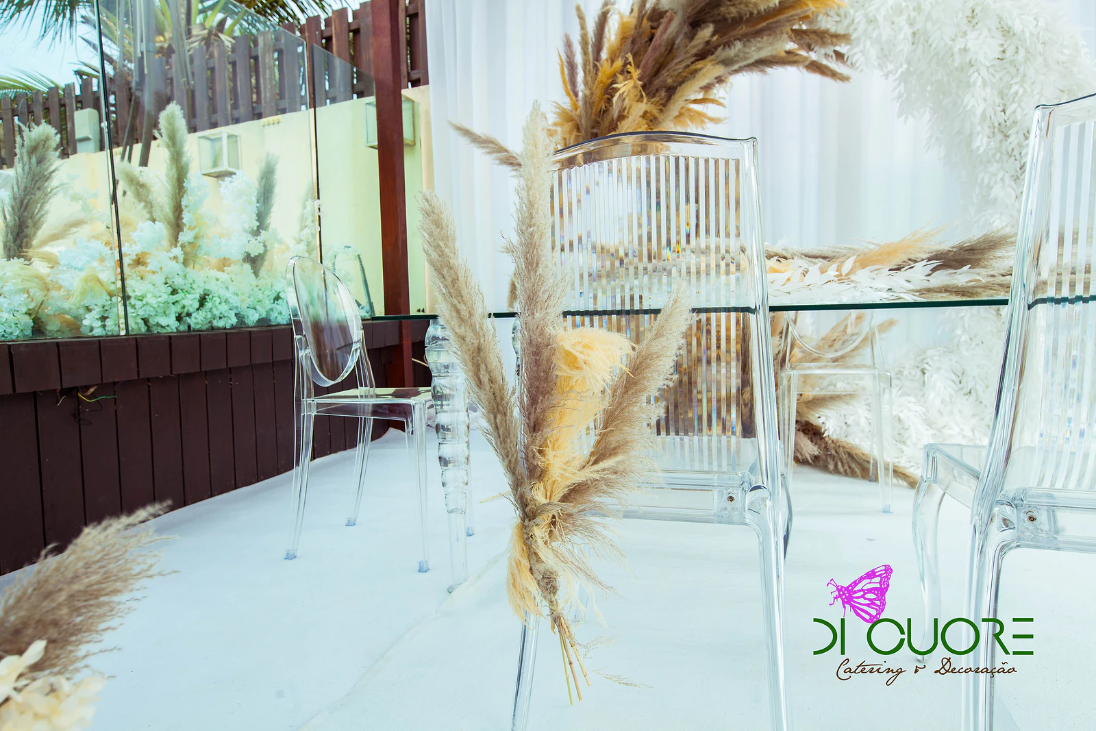 A bunch of pampas grass tied to a clear chair, beside the glass ceremony table