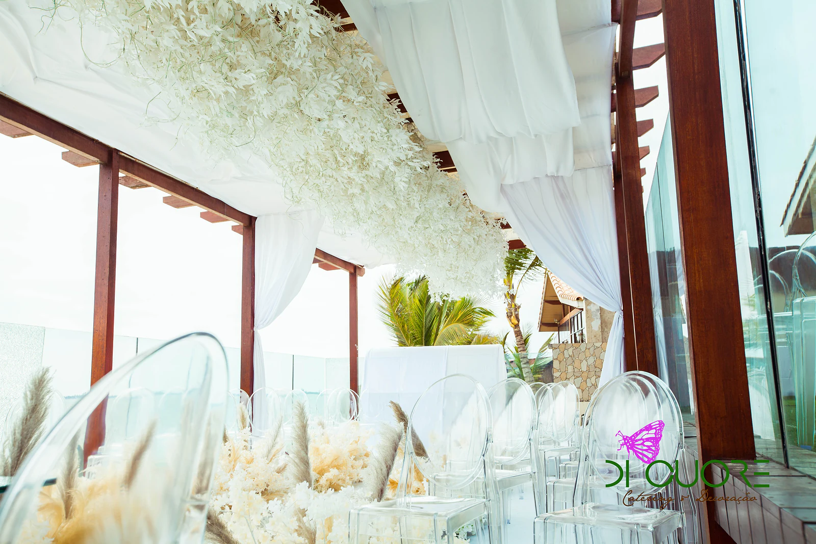 White aisle with clear chairs on one side and branches of white flowers and pampas on the other, leading towards the sea and the far end of the ceremony