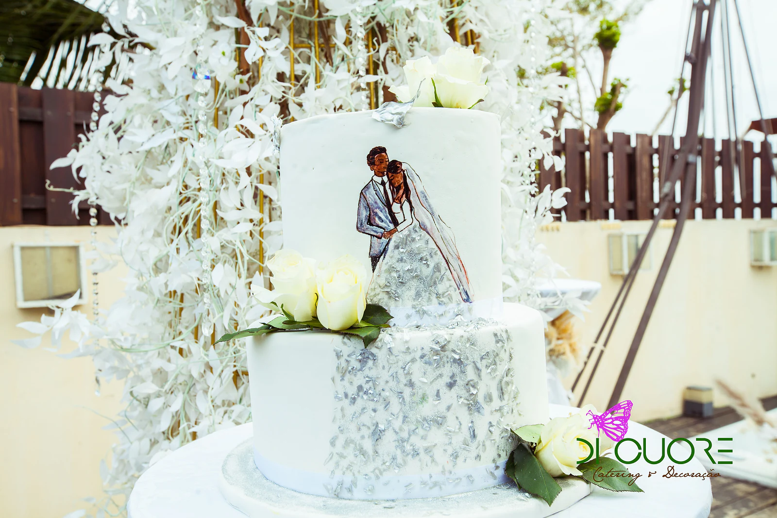 Two-tier white cake with silvery details, white roses and the drawing of a couple, in front of a curtain of white leaves