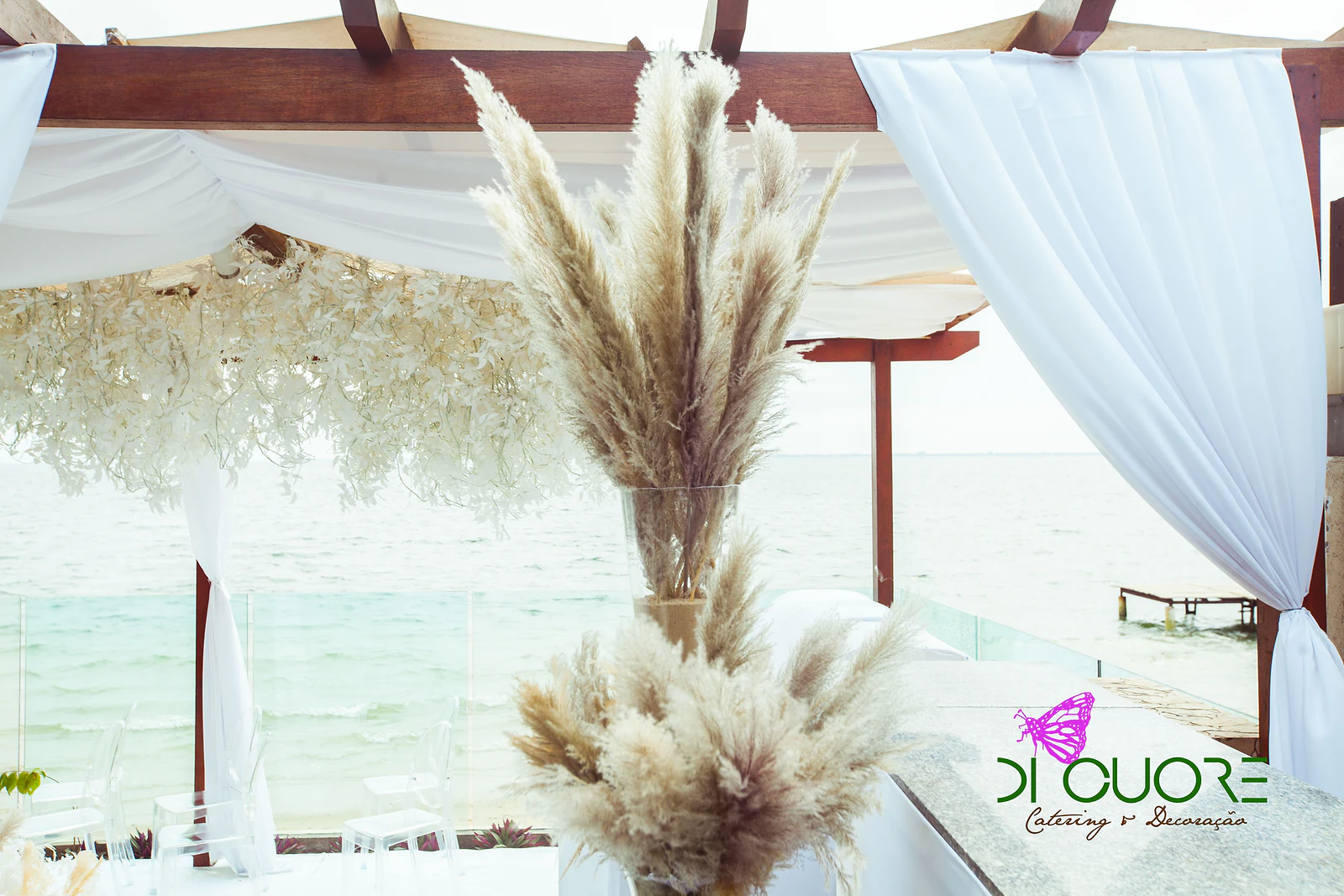 Tall plumes of pampas grass in glass vases, in front of the white-draped pergola and the sea