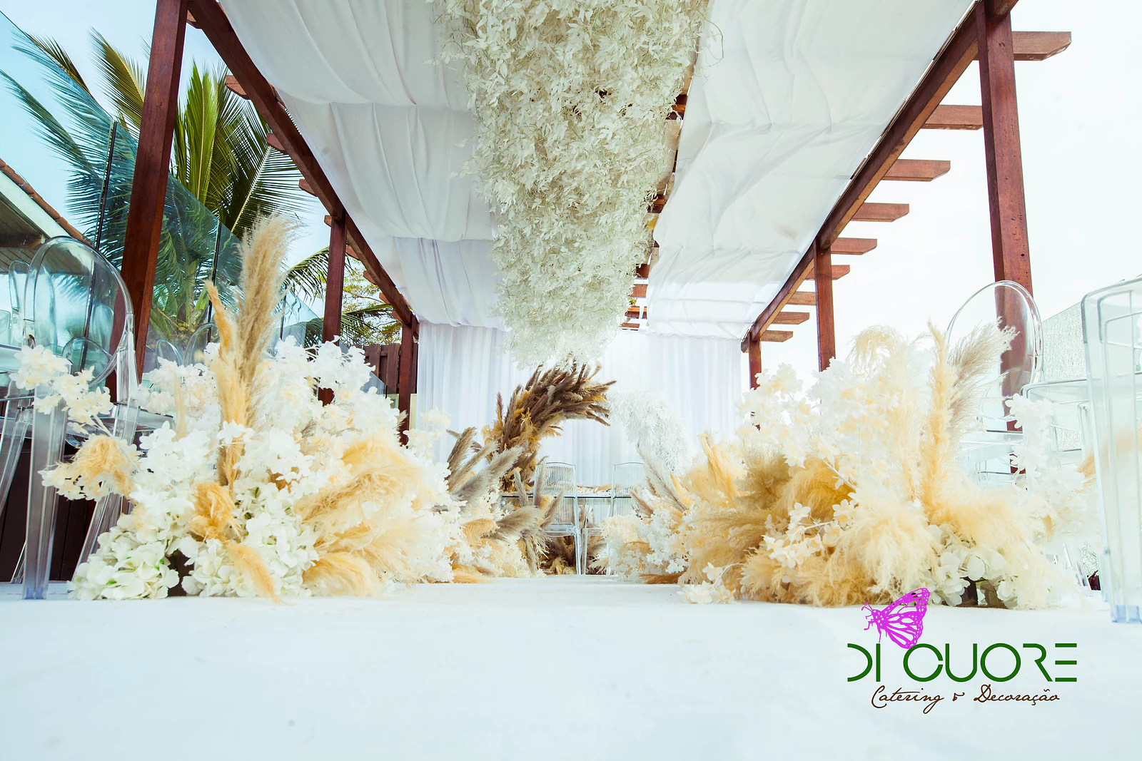 Low arrangements of white flowers and pampas grass along the white aisle, beneath the ceiling of hanging leaves