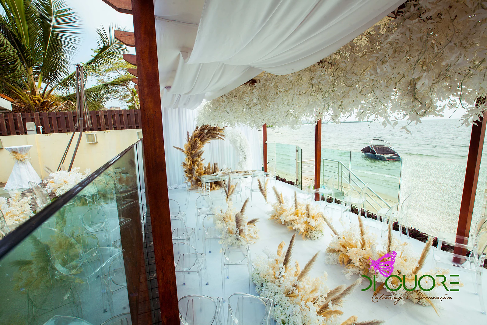 Angled view of the aisle with pampas and white flowers, clear chairs and the arch at the far end, beneath the ceiling of white leaves, with the sea to the right
