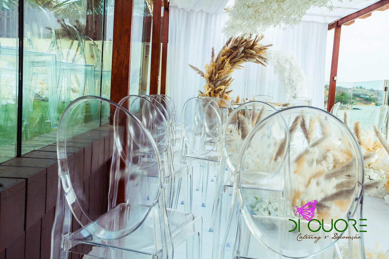 A row of clear chairs with plumes of pampas grass between their backs, beside a glass wall