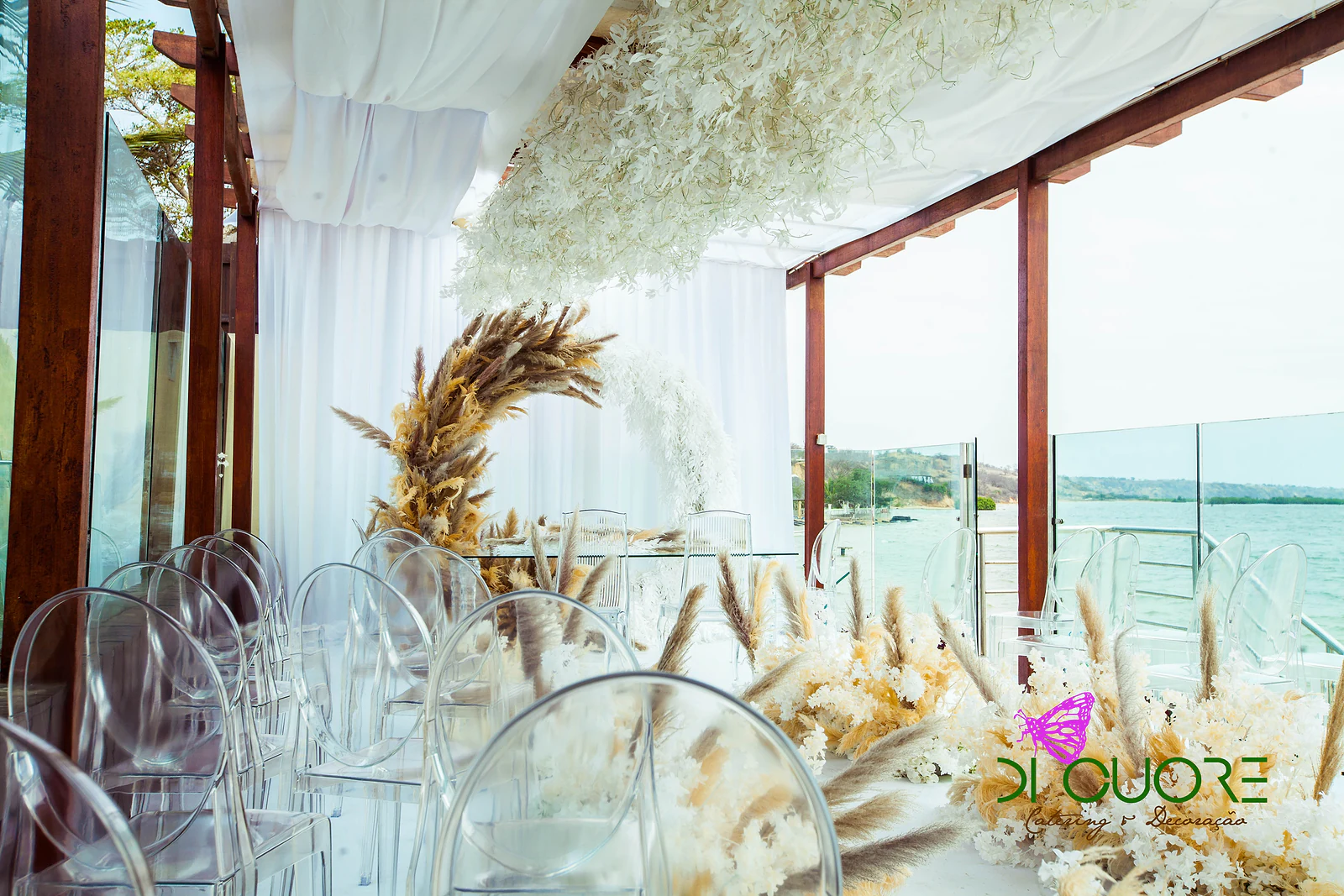 A row of clear chairs and pampas-grass arrangements beside the ceremony arch, with the sea beyond